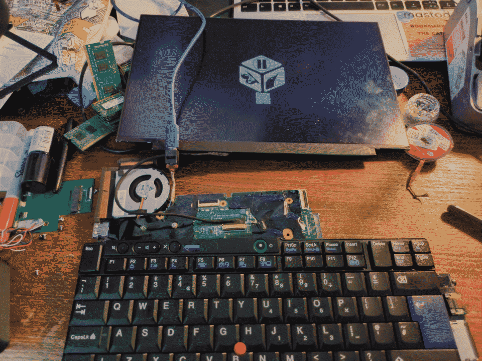 The motherboard and screen are connected with the motherboard just laying ontop. The screen is on with the heads logo displaying. The desk is messy with lots of random computer parts along with nail polish and sharpie